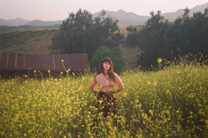 A person stands in a field of tall yellow flowers, with a background of trees and mountains under a hazy sky.