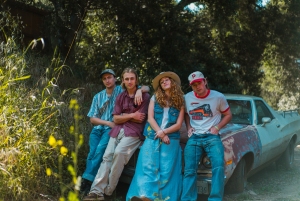 Four people leaning against an old car in a wooded area, wearing casual clothing and hats, with trees and grass in the background.