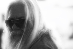 A person with long white hair, a beard, and sunglasses is facing sideways in a black and white image.