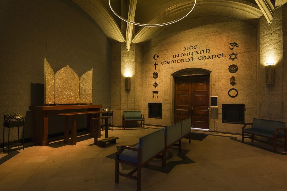 Interior of an interfaith chapel with chairs, a wooden altar, and various religious symbols on the wall beside a wooden door.