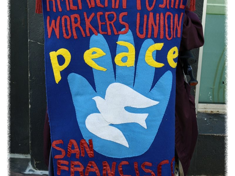 A person holds a banner reading "American Postal Workers Union Peace San Francisco Local" with a blue hand and white dove design.