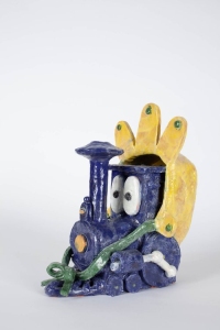A blue clay figure resembling a train with large eyes and a yellow crown-like structure on top sits against a plain background. It has a green bow at the front.