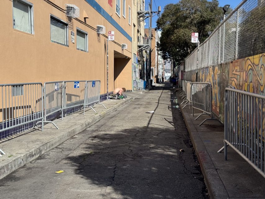 A narrow urban alley with metal barriers along left and a colorful mural on the right. A person is seated on the ground near a building on a sunny day.