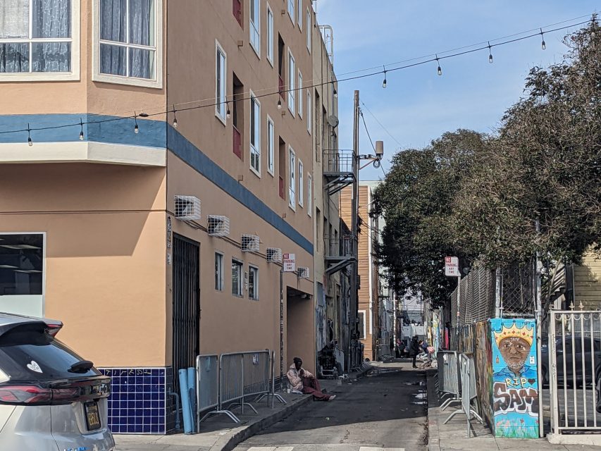 An urban alley with a person sitting on the ground near a building, a painted mural on a fence, and string lights above.
