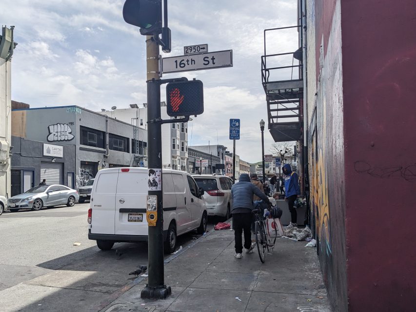Street scene at 16th St. A pedestrian crosses by with a bicycle, with buildings and graffiti in the background. Traffic signal shows "Don't Walk.