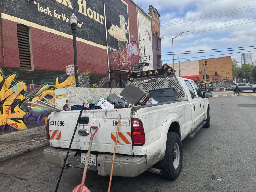 White truck filled with debris and cleaning tools parked on a graffiti-covered street. Nearby landmarks include a mural and urban buildings.