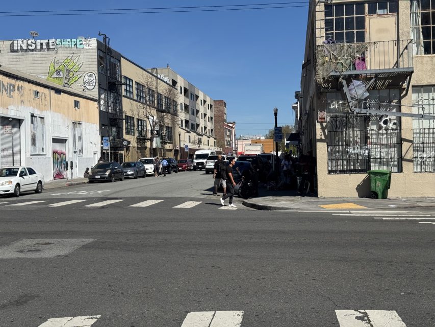 Urban street scene with people walking, cars parked along the road, and graffiti on buildings. Crosswalk markings are visible on the pavement. Bright sunny day.