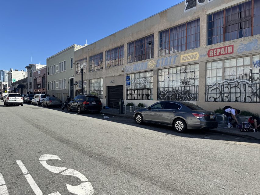 Street view of an industrial area with graffiti on buildings, parked cars, and a few people near a building. Sunny day with a clear blue sky.