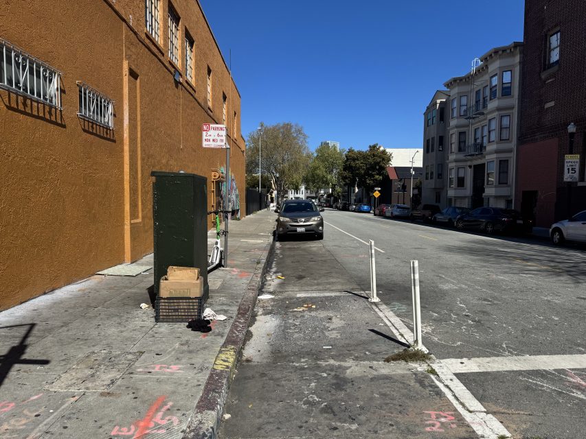 Urban street scene with a parked car beside a sidewalk. Buildings line both sides, and a "No Parking" sign is visible. The sky is clear and blue.
