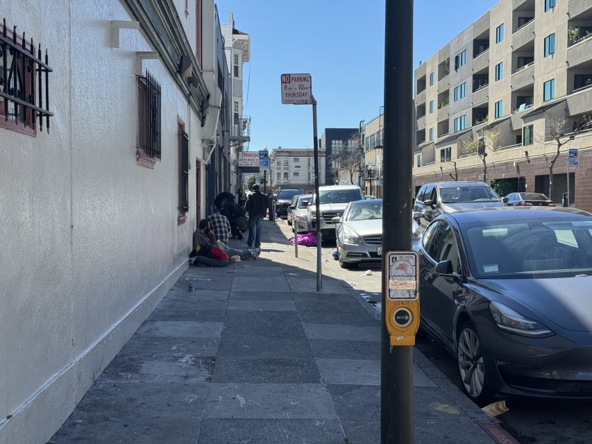 Street view with parked cars, a "No Parking" sign, and a group of people sitting on the sidewalk near a building. A pedestrian crossing button is visible in the foreground.