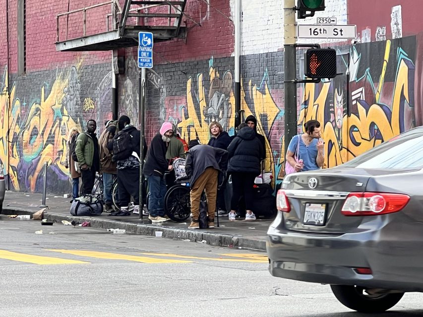 A group of people gathers near a vibrant graffiti-covered wall at the corner of 16th St. Plaza, close to a bustling crosswalk and traffic light. A car is partially visible in the foreground, adding to the lively city scene.