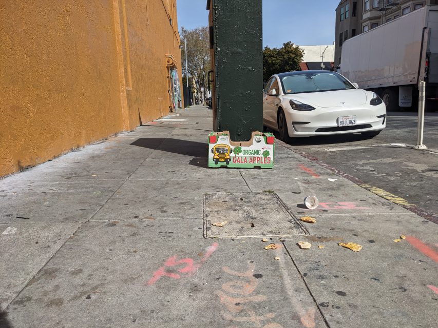 An empty organic Gala apples box on a city sidewalk, surrounded by litter. A white car is parked nearby.