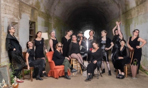 A group of people dressed in black and gray clothing pose inside a tunnel. They sit and stand around two chairs, with varied poses and expressions.