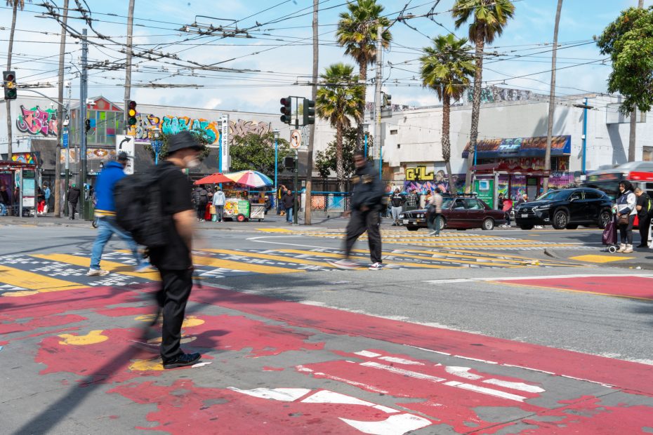 People walking at a busy urban intersection with colorful crosswalks, palm trees, and graffiti-covered buildings in the background.