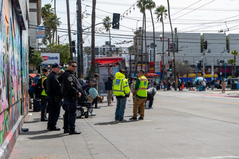 Police and workers in yellow vests stand on a city sidewalk near a colorful mural, with pedestrians and palm trees in the background.