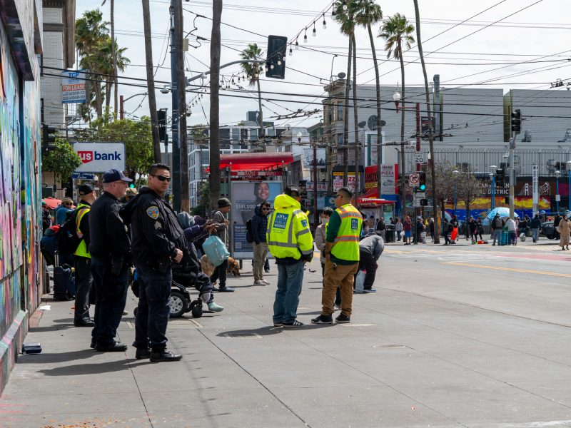 Police and workers in yellow vests stand on a city sidewalk near a colorful mural, with pedestrians and palm trees in the background.
