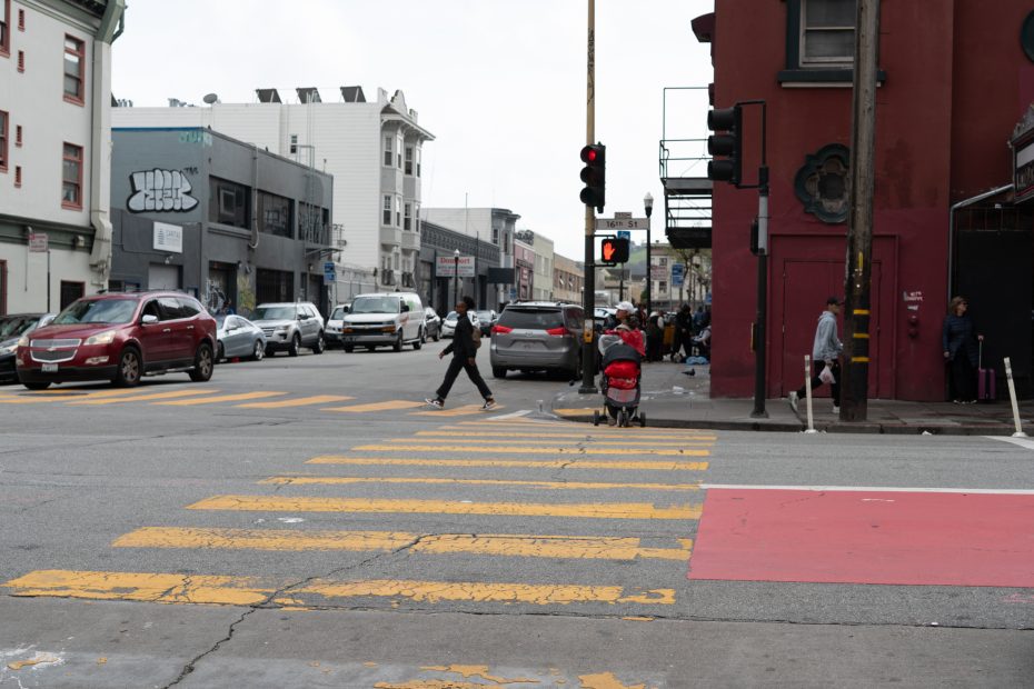 Urban street scene with cars waiting at a red light, a person pushing a stroller on a crosswalk, and pedestrians on the sidewalk. Buildings line the street under a cloudy sky.