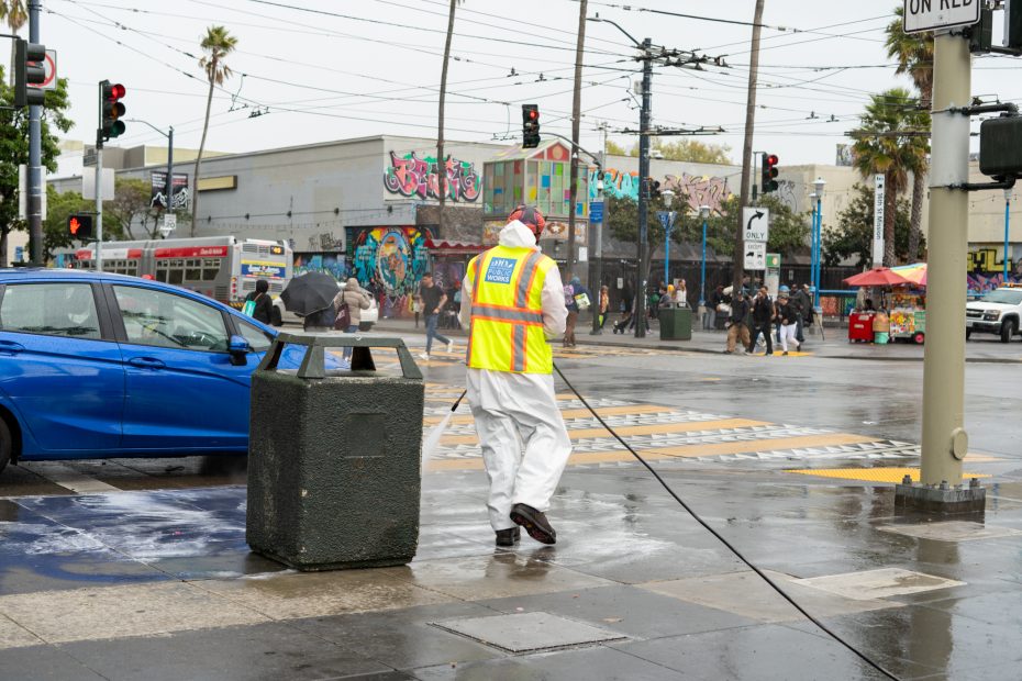 A person in a high-visibility vest and protective clothing power-washes a sidewalk on a rainy day in an urban area. Traffic and pedestrians are visible in the background.