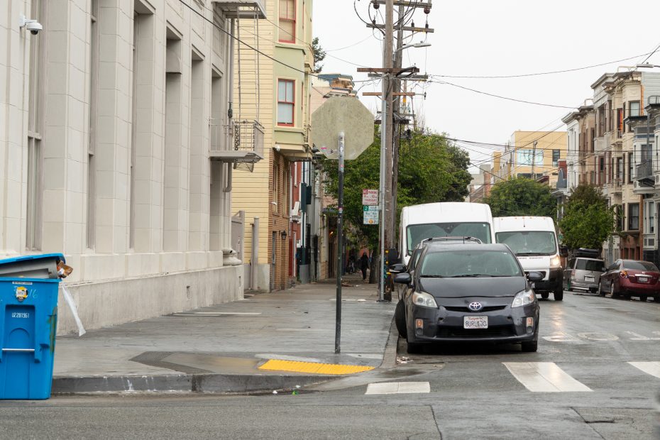 Street scene with parked cars, a blue recycling bin on the sidewalk, and multiple buildings. Power lines and a stop sign are visible. Overcast weather.