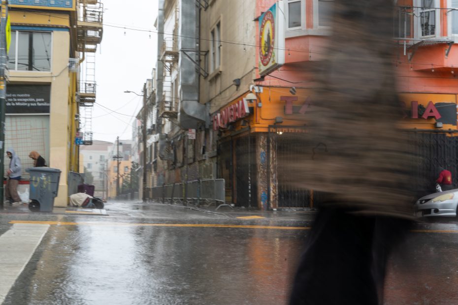 Blurred pedestrian walking on a rainy street. Buildings and shops, including a taqueria, line the wet pavement. A few people are visible along the sidewalk in the background.