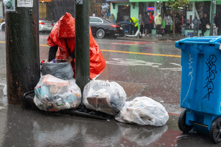 Person in an orange poncho by garbage bags on a rainy street.