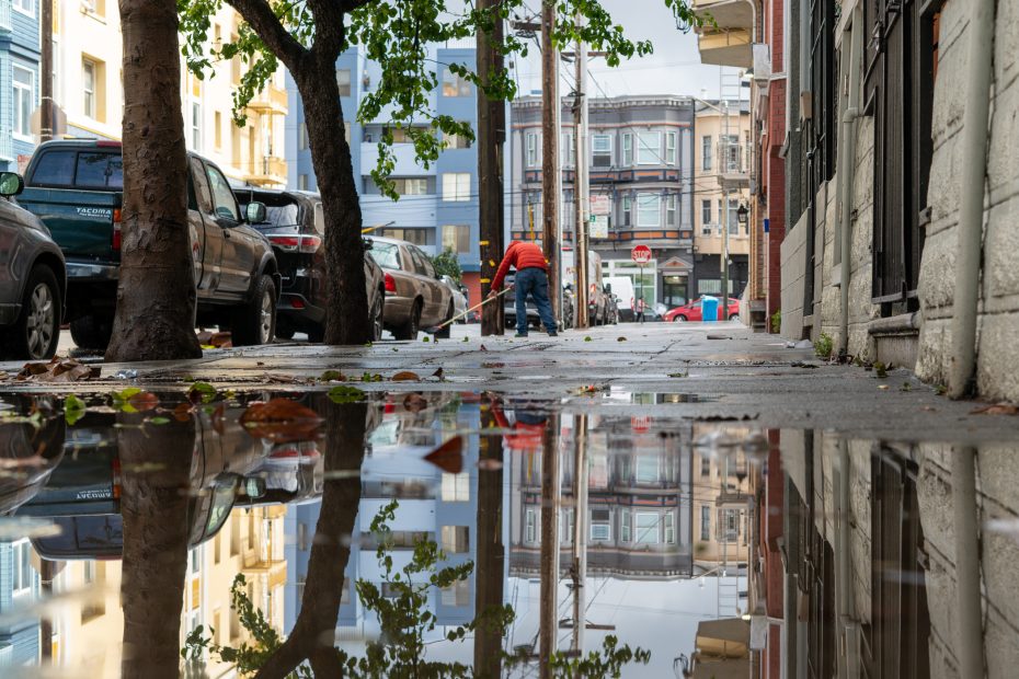 A person sweeps the sidewalk near a tree-lined street with cars parked along the curb. Buildings are reflected in a large puddle in the foreground.