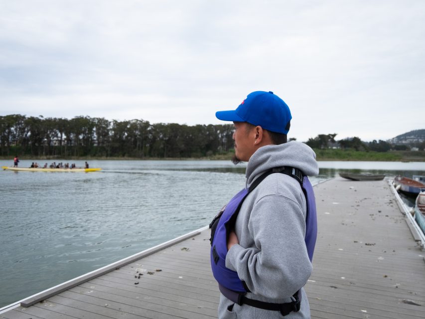 A person wearing a blue cap and gray hoodie stands on a dock, observing a rowing team on the water in the distance.