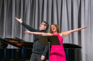 Two people stand in front of a grand piano with arms outstretched, singing passionately. The man is in dark clothing and the woman is in a bright pink dress. They are both smiling.