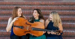 Three women sitting on steps with musical instruments, including a cello, smiling and interacting with each other.