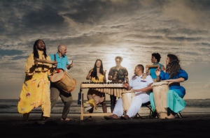 A group of people on a beach play drums and a wooden xylophone under a cloudy sky with the sun behind them.