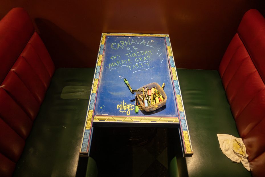 A table with a blue top displays chalk-written notes about Mardi Gras and Carnaval. A container with chalk pieces is on the table, surrounded by green and red booth seating.