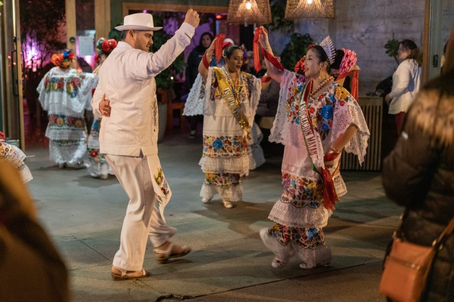 A man in white dances with women in colorful traditional dresses adorned with flowers, inside a lively, warmly lit venue.
