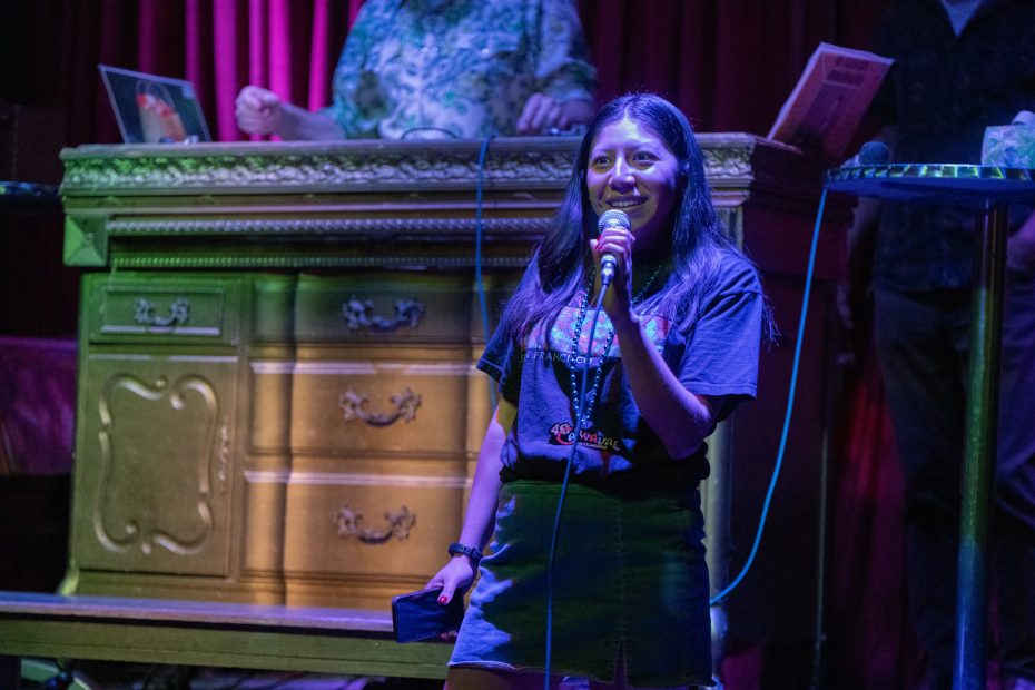 Person holding a microphone, standing in front of a wooden cabinet with a purple curtain backdrop.