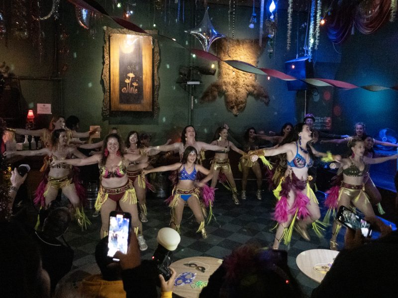 A group of dancers in vibrant costumes performs a choreographed routine on a dimly lit stage, with audience members capturing the moment on their phones.