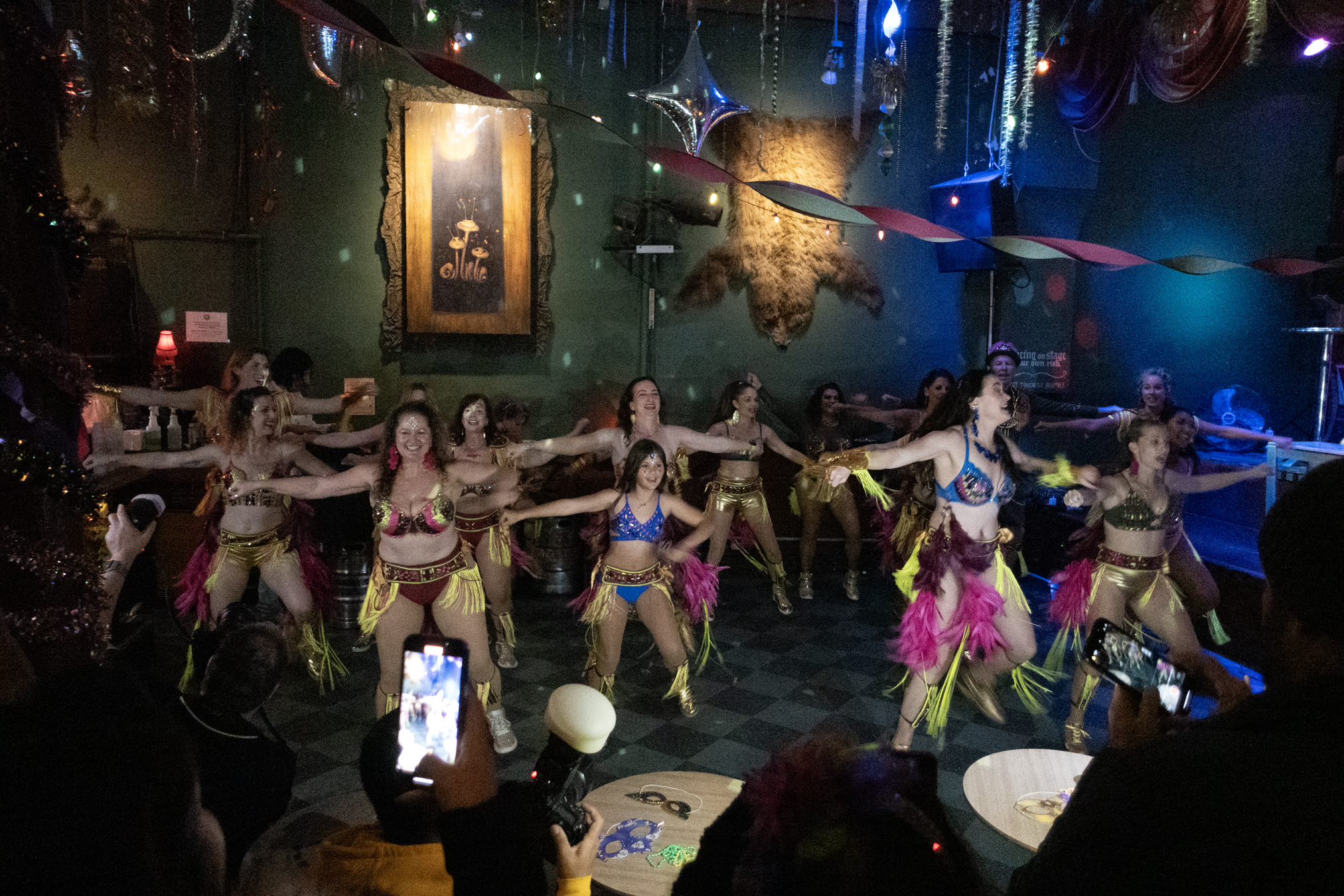A group of dancers in vibrant costumes performs a choreographed routine on a dimly lit stage, with audience members capturing the moment on their phones.