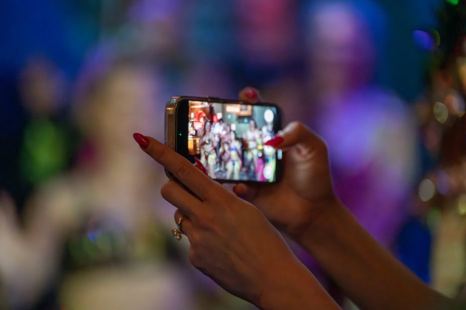 Person holding a smartphone horizontally, taking a photo of a group. Background is blurred with colorful lights.