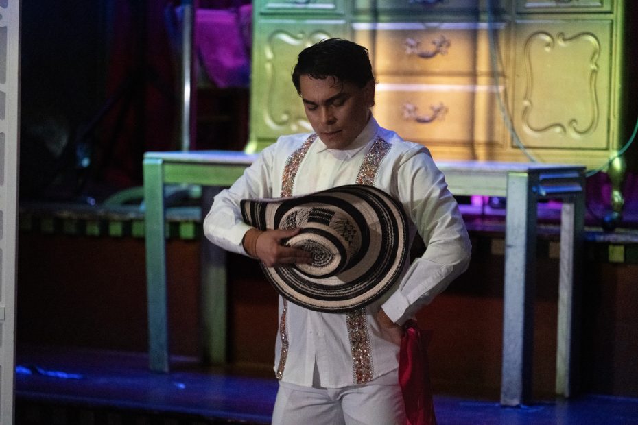 A person in a white outfit holds a black and white striped hat in a performance setting, standing in front of an ornate background.