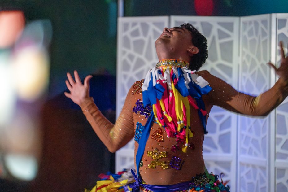 Person wearing a colorful, patterned costume with raised arms and tilted head, performing expressively in a dimly lit setting.