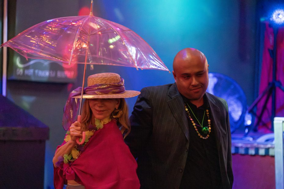 Two people standing under colorful lighting; one holds a clear umbrella and wears a straw hat with a purple scarf and pink wrap, while the other wears a striped suit and beaded necklaces.