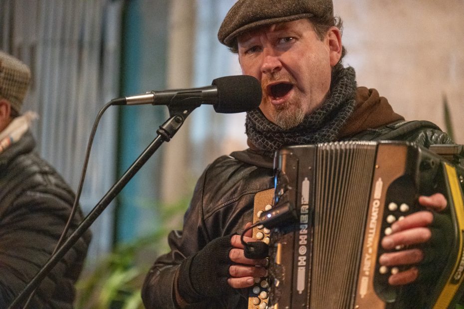 A man wearing a hat and scarf sings into a microphone while playing an accordion.