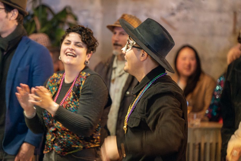 Two people wearing festive costumes and masks, smiling and clapping in a lively indoor setting. Other people are visible in the background.
