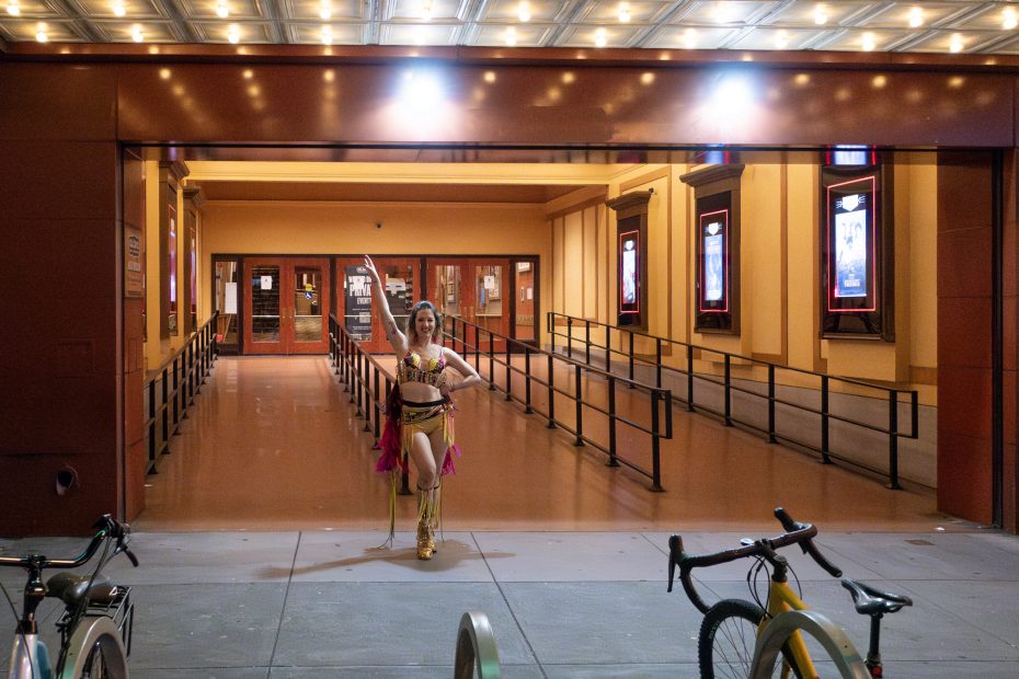 A person in a belly dance outfit poses outside a theater entrance. Two bicycles are parked in the foreground.