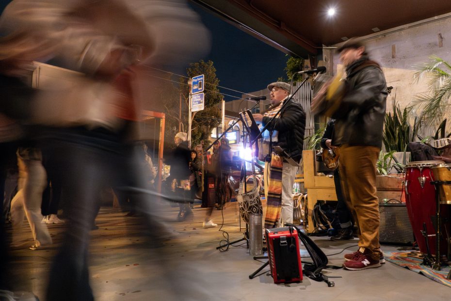 Street musicians perform at night with blurred pedestrians walking by. A guitarist and a wind instrument player are visible, surrounded by musical equipment.