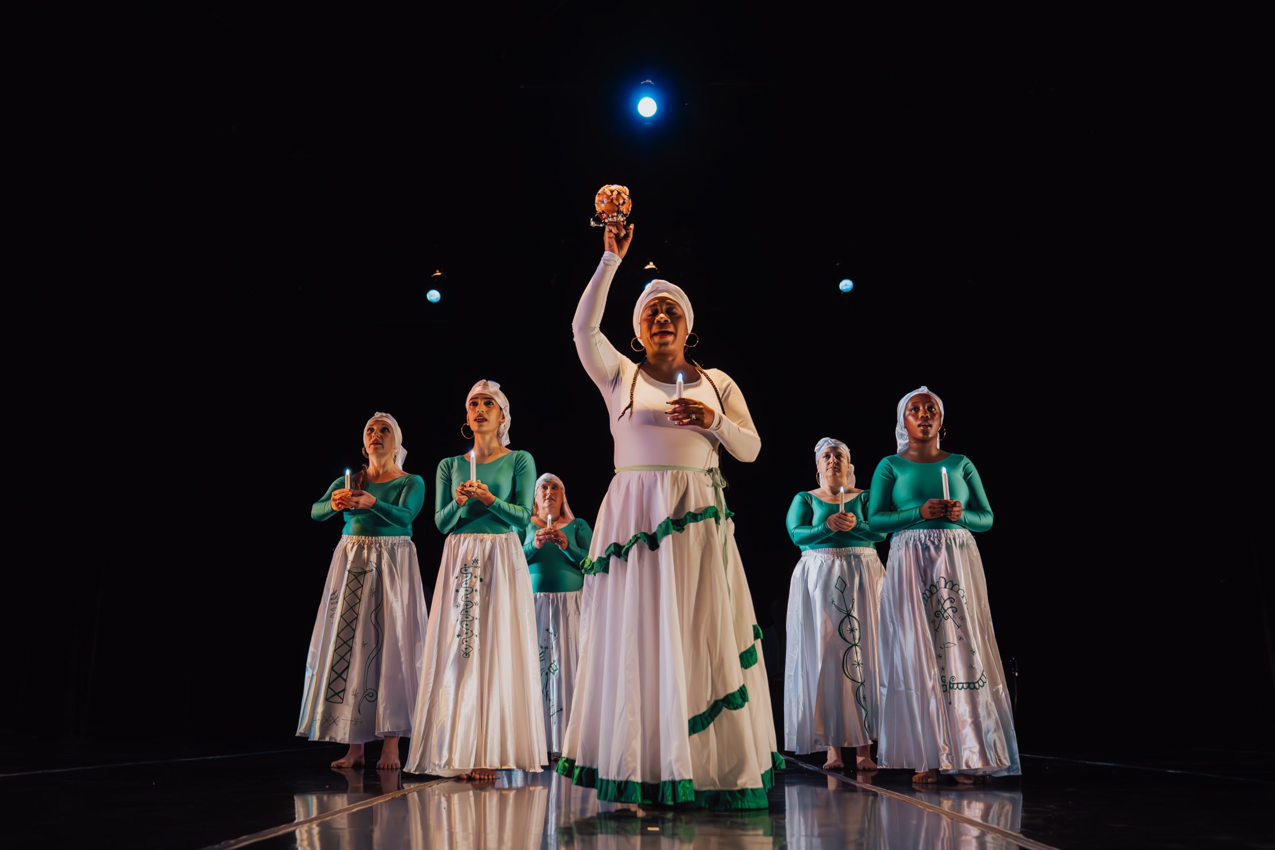 Seven people in white and green outfits perform on stage under spotlights, with one person holding an object up.