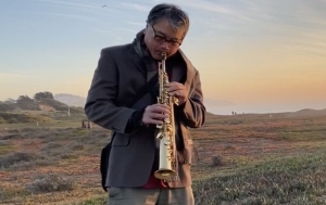 A person plays a saxophone outdoors in a grassy area at sunset, wearing a jacket and glasses.