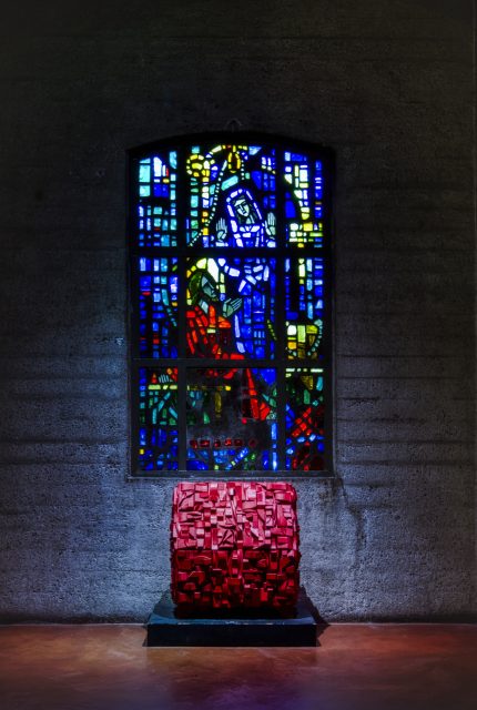 A colorful stained glass window with an abstract design, featuring blues, reds, and yellows. Below it sits a red-textured cube on a pedestal against a dark wall.