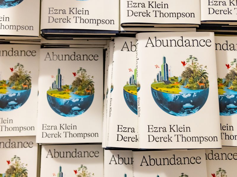 A stack of books titled "Abundance" by Ezra Klein and Derek Thompson, featuring an illustrated cover with a cityscape and greenery on a globe.