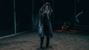Person stands with arms raised in a dimly lit room with sand on the floor, while another person sits in the background.