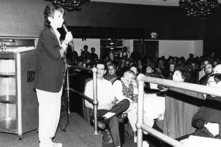A person speaks into a microphone on stage in a venue. The audience, seated at tables, listens attentively. The image is in black and white.