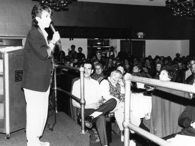 A person speaks into a microphone on stage in a venue. The audience, seated at tables, listens attentively. The image is in black and white.
